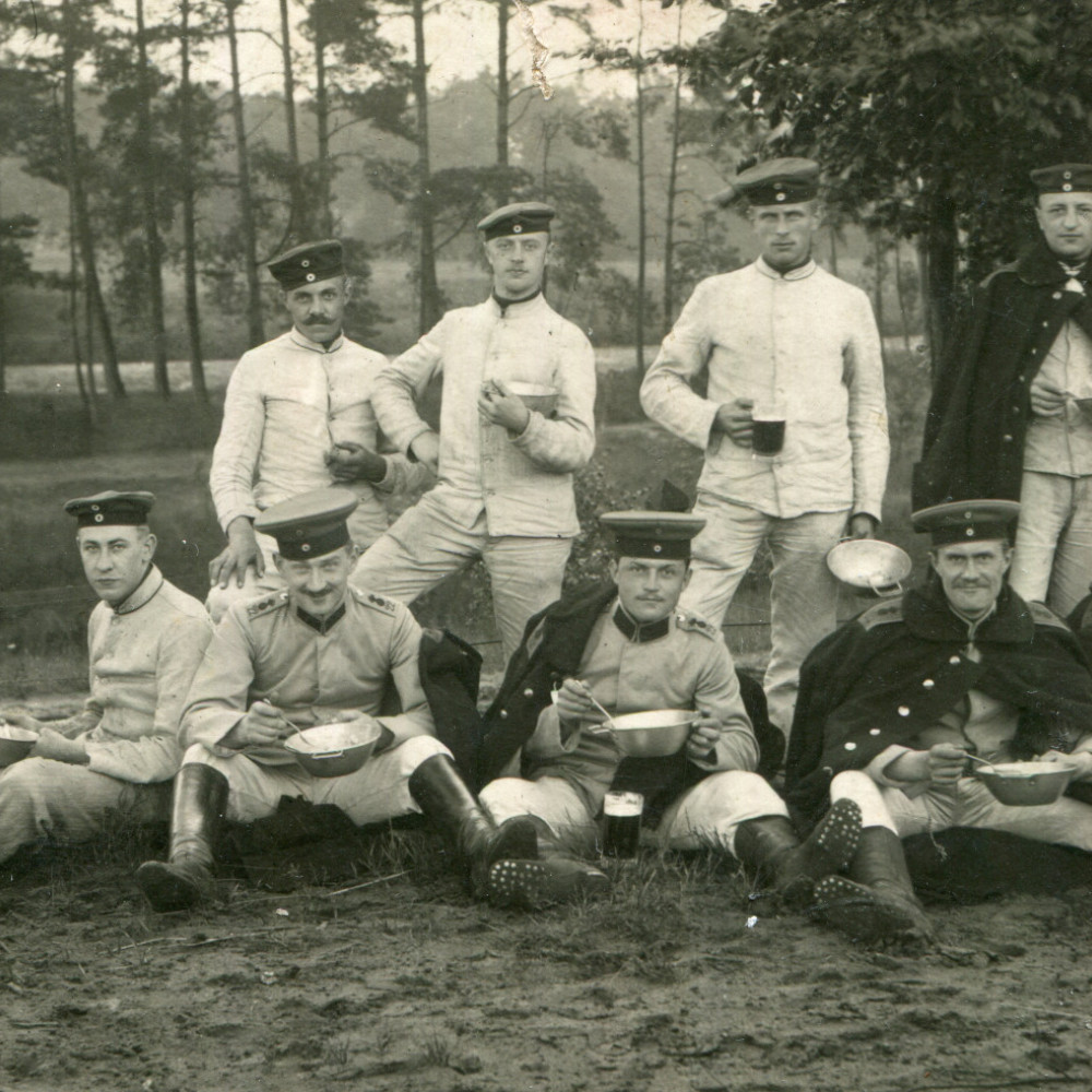 Angehörige des FAR 22 bei der Mahlzeit wohl im Rahmen einer Truppenübung ggf. im Sennellager. Die Soldaten tragen teilweise Drillich und teilweise Litewkas bzw. umgehängte Mäntel. Gut zu sehen sind die genagelten Infanteriestiefel.